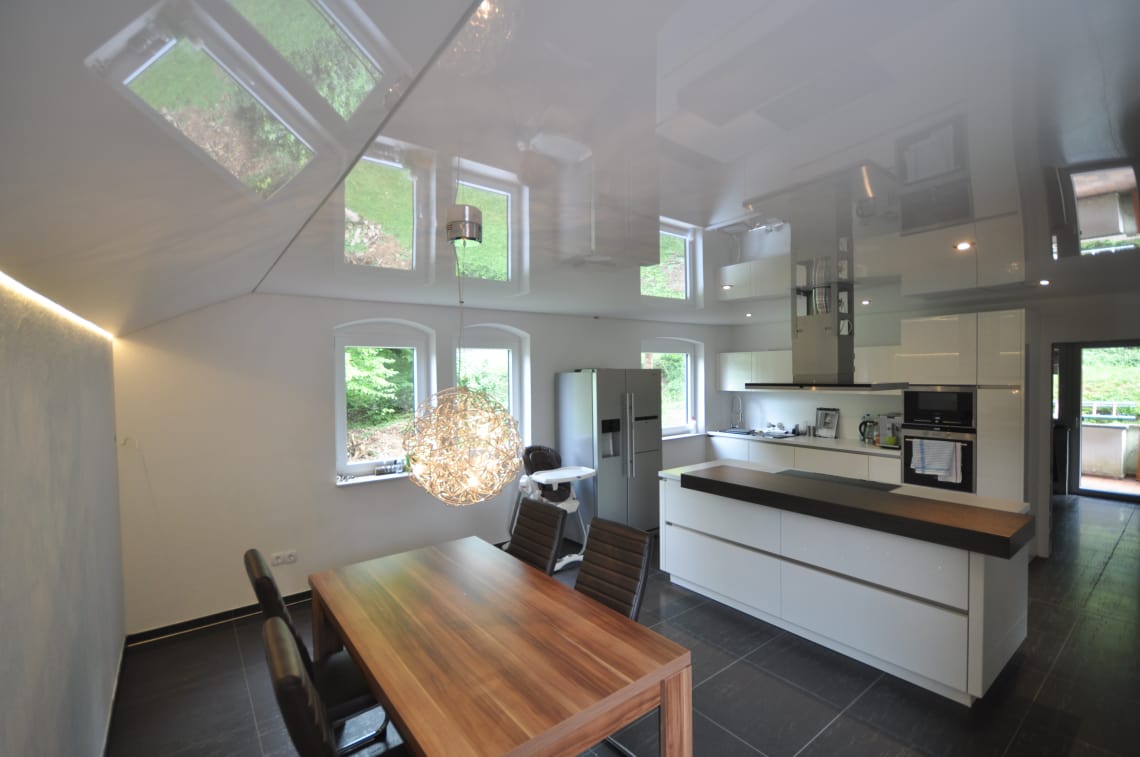 Lackspanndecke in der Küche mit Dachschräge in weiss Hochglanz, Beleuchtung mit LED-Einbauleuchten, Penelleuchte über dem Tisch und LED-Lichtkanal in der Schattenfuge